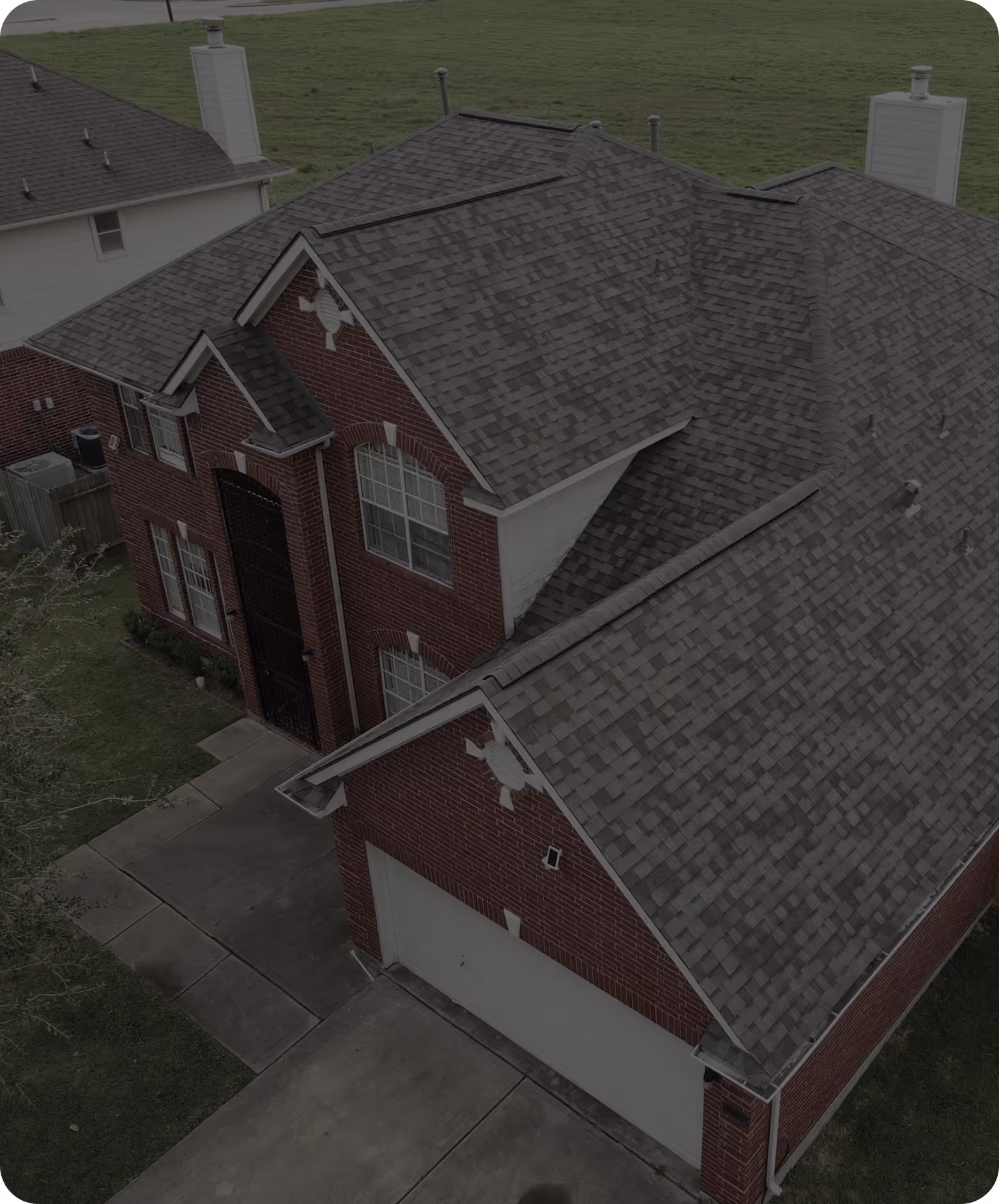 Aerial view of a red brick two-story house with a gray shingle roof, driveway, and green lawn.
