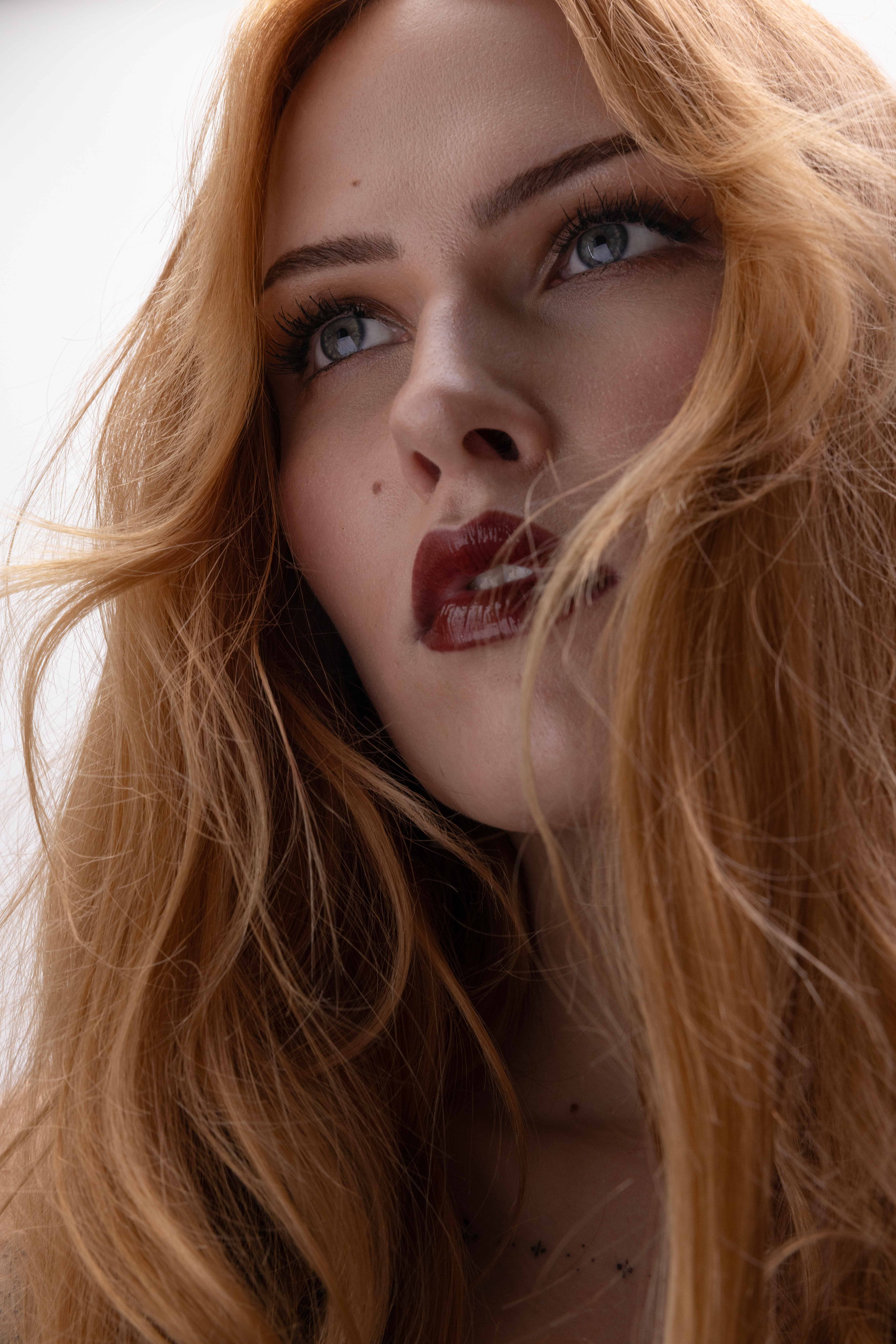 Artistic portrait of a woman with vibrant red crimped hair, soft pink blush, and a serene expression on a pastel gradient background – beauty photography by Kendra Storm Rae in Berlin.
