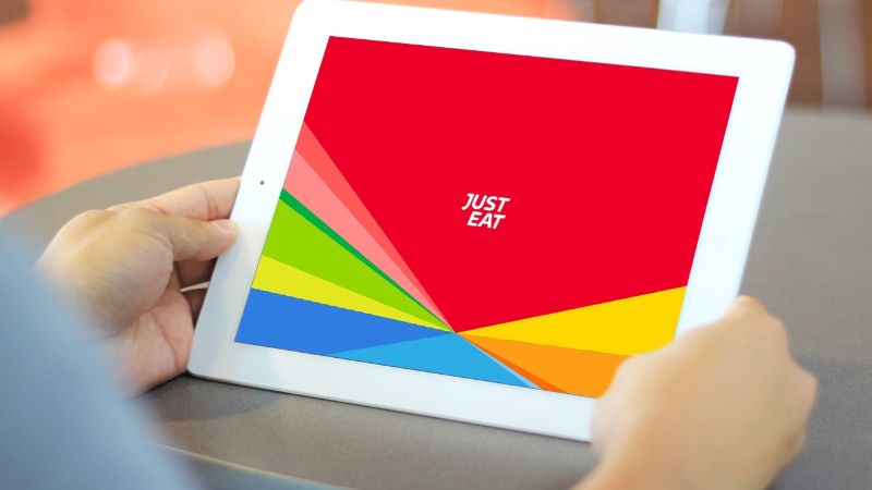 Hands holding a white tablet displaying a colorful screen with the text 'JUST EAT' in the center.