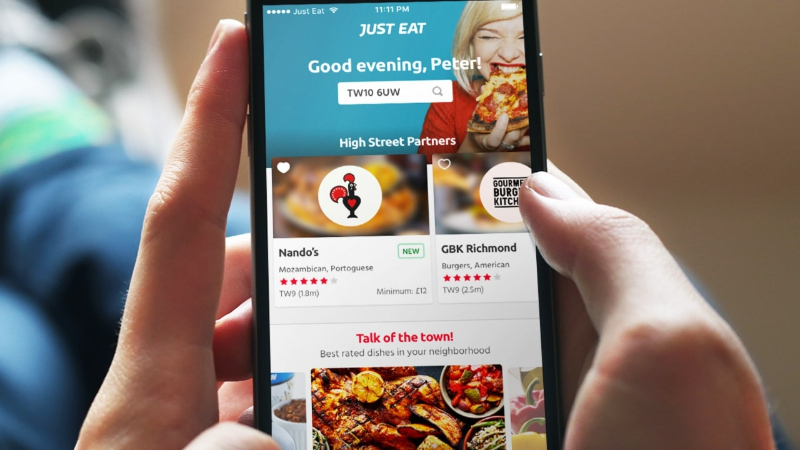 Person holding phone displaying Just Eat app with restaurant options and a food photo.