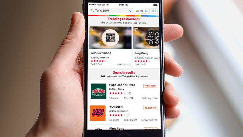 Hand holding phone showing Just Eat app with trending restaurants and search results for TW10 6UW.