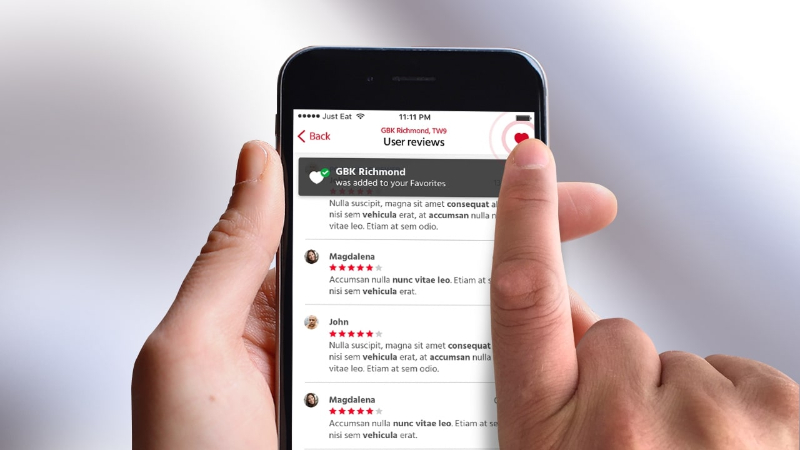 Hand holding smartphone showing user reviews and adding GBK Richmond to favorites with a heart icon.