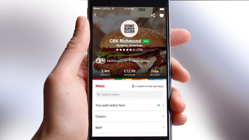 Hand holding a phone displaying Gourmet Burger Kitchen app menu with burger image and options.