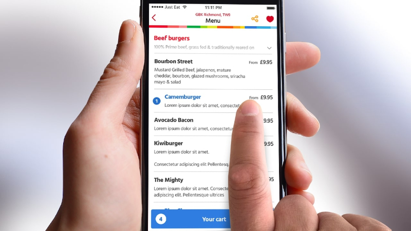 Hand holding a smartphone showing a menu screen with beef burgers and prices from a food app.