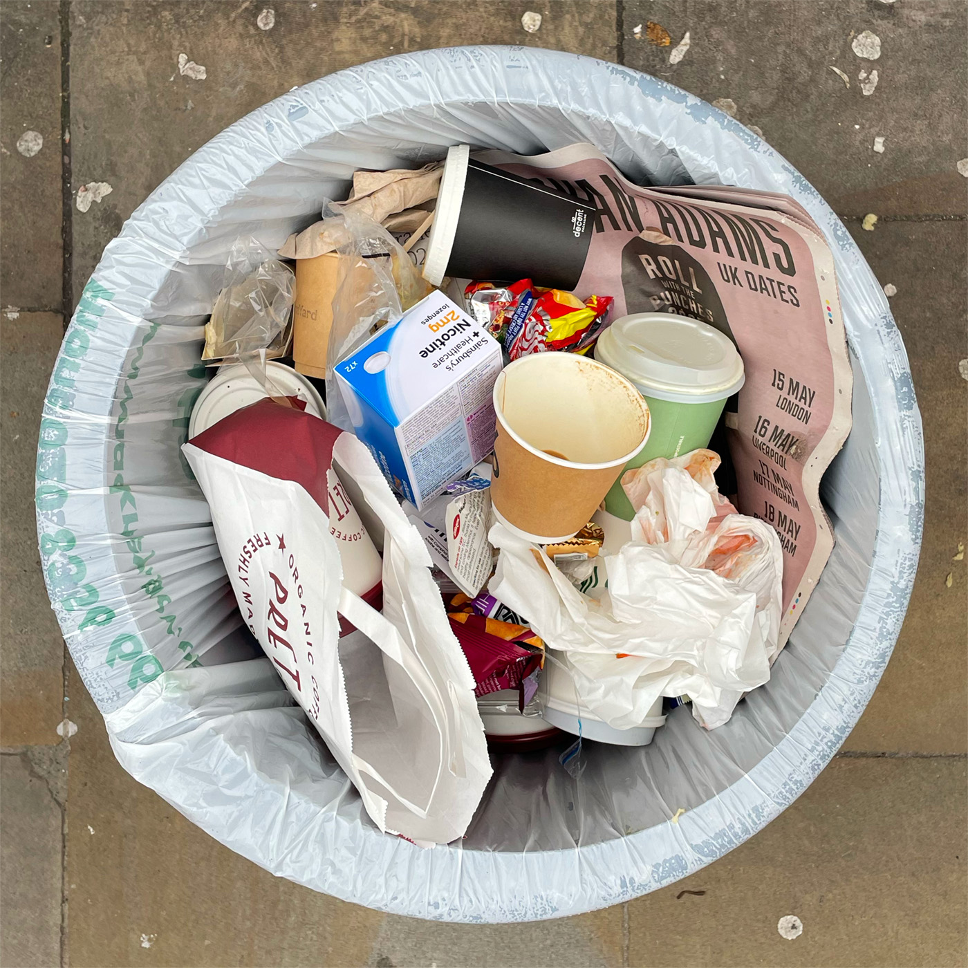 An open rubbish bin on the pavement filled with paper cups and newspaper.