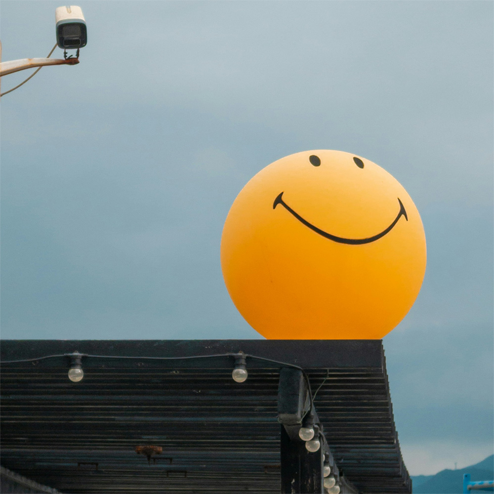 a large yellow balloon with smiling face perched on the edge of a corrugated metal roof being watched by a cctv camera