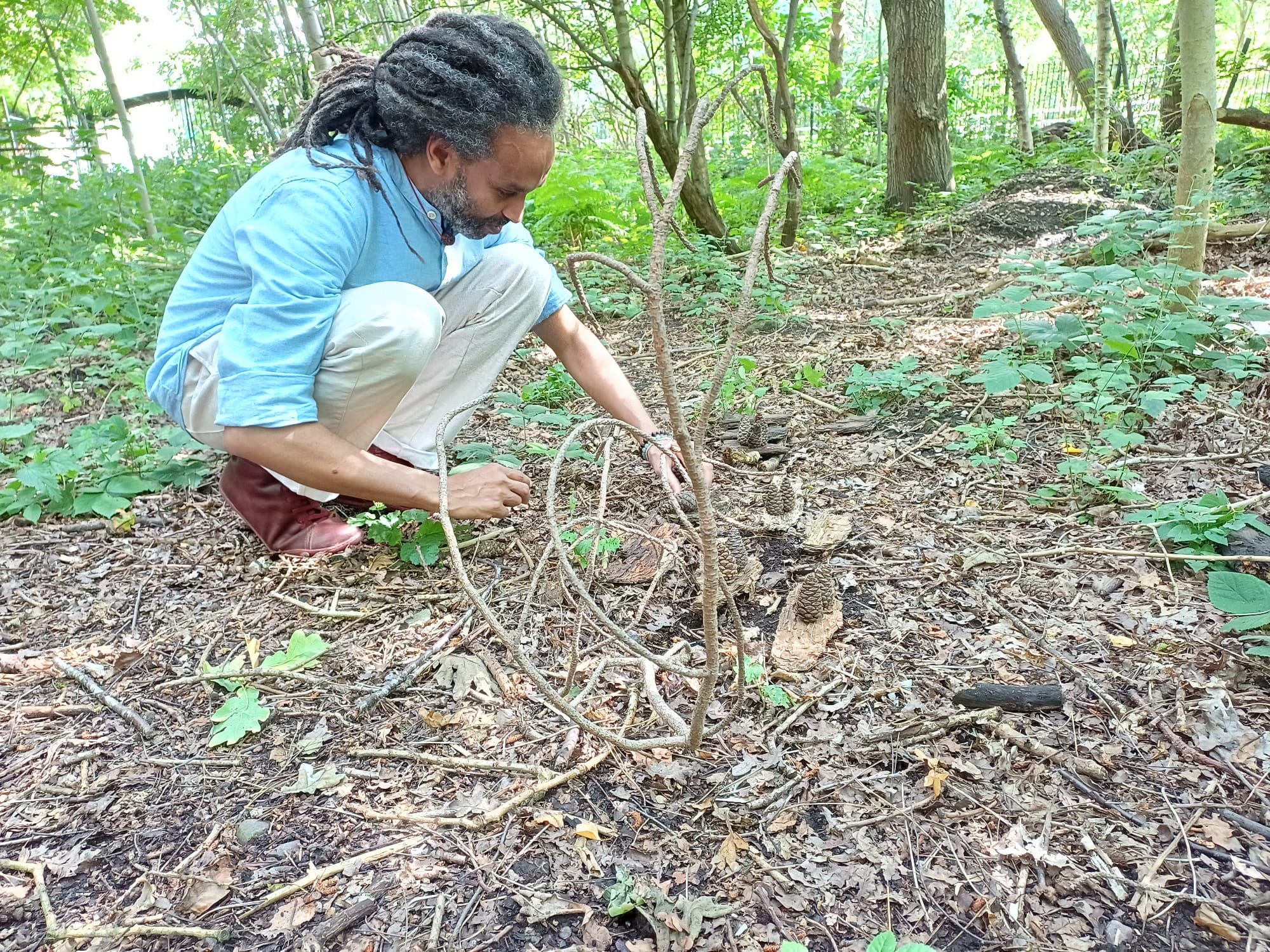 A person with dark skin and long tied back hair, blue short sleeved shirt, white trousers and sandals is crouched down on a forest floor. The ground around is brown with woody and  leafy vegaetation around and he is making some kind of land art sculpture. All around is green leafy vegetation and trees in full leaf.