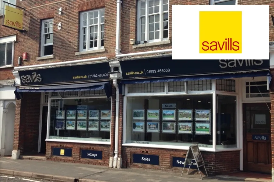 Street view of a brick Savills real estate office with display windows showcasing property listings under awnings labeled Lettings, Sales, and New Homes.
