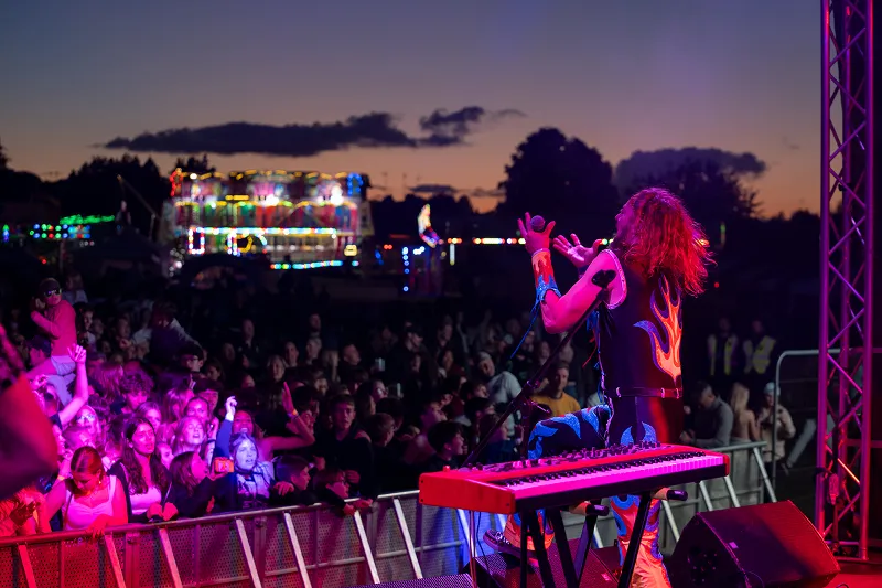 Singer with flame-patterned outfit passionately performing on stage at dusk in front of a large crowd and illuminated carnival rides.