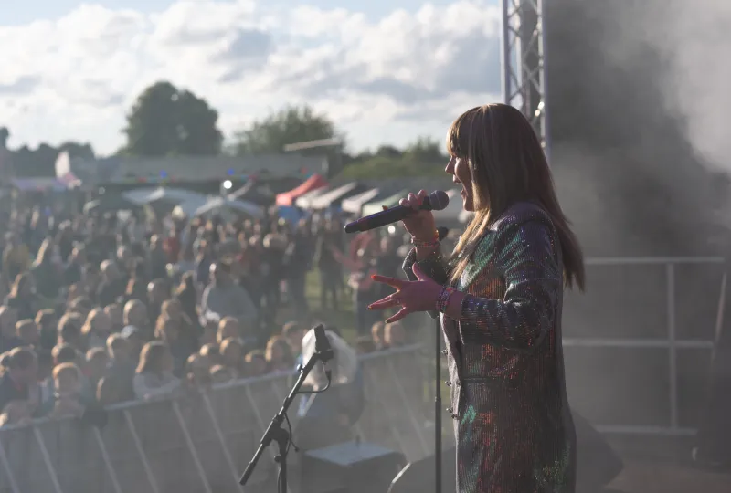 Female singer performing on stage at an outdoor concert in front of a large crowd during daylight.