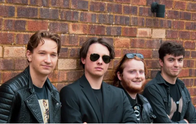 Four young men standing against a brick wall, dressed casually with one wearing sunglasses and another with glasses on his head.