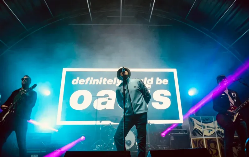 Three musicians performing on stage with bright blue and purple lights and a large sign behind reading 'definitely could be Oasis'.
