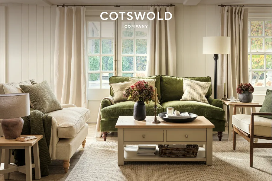 Cozy living room with beige and green sofas, wooden coffee table with flowers, and large windows with cream curtains.