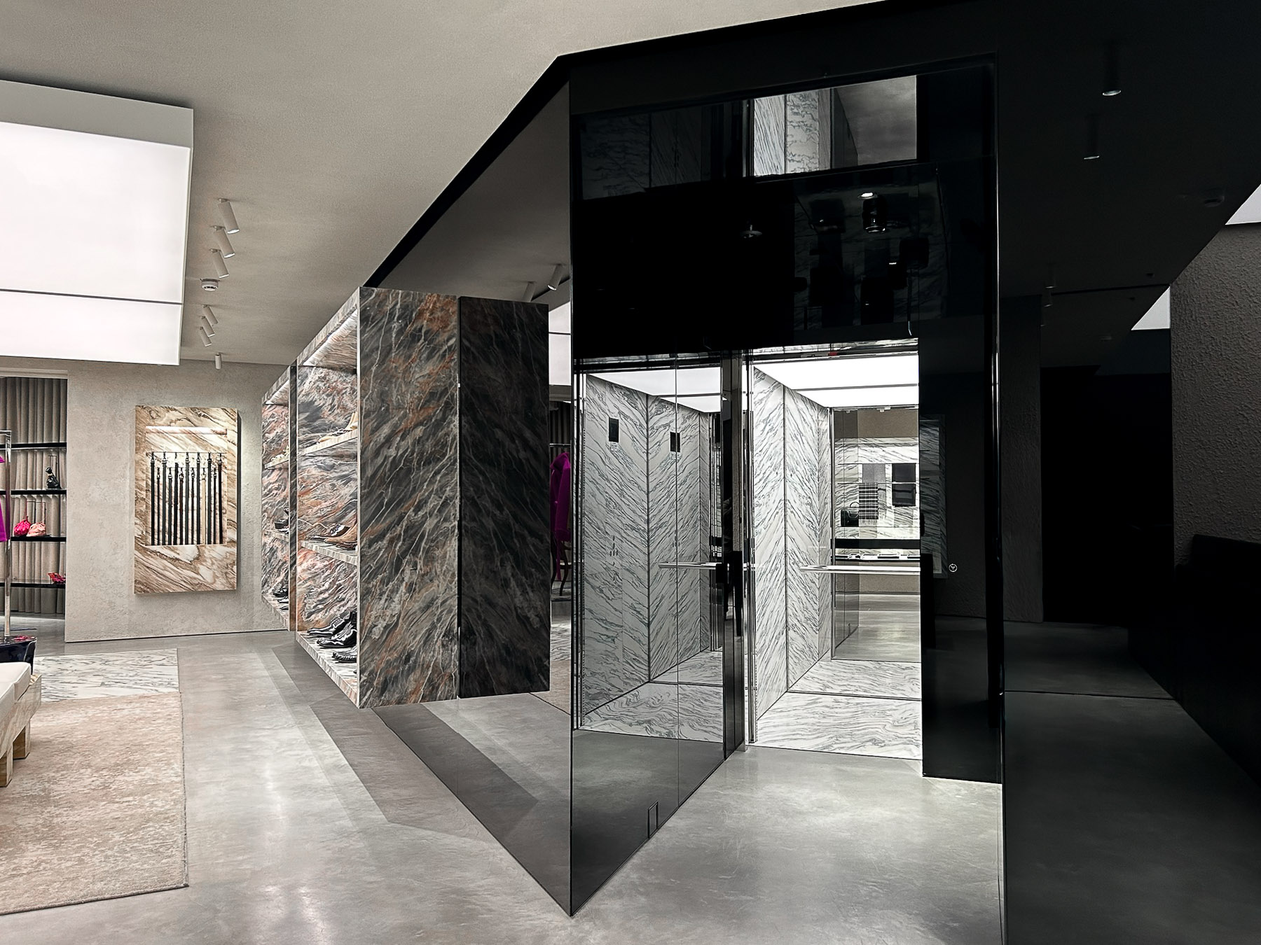 Modern retail store interior with polished concrete floor, marble wall panels, black glass elevator, and shelves displaying shoes and handbags.