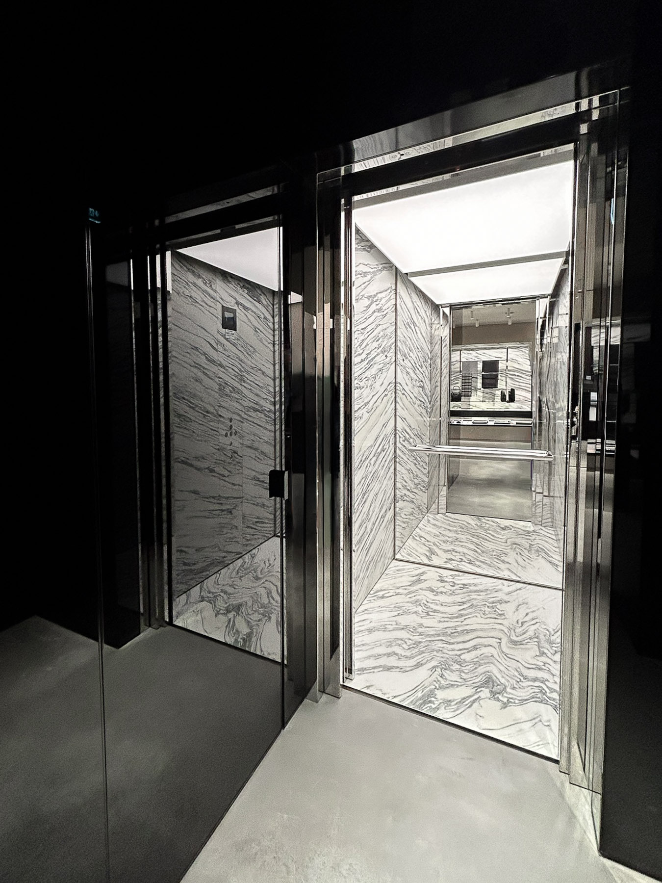 Modern elevator interior with white and gray marbled walls and floor, illuminated ceiling, and reflective metal frame.
