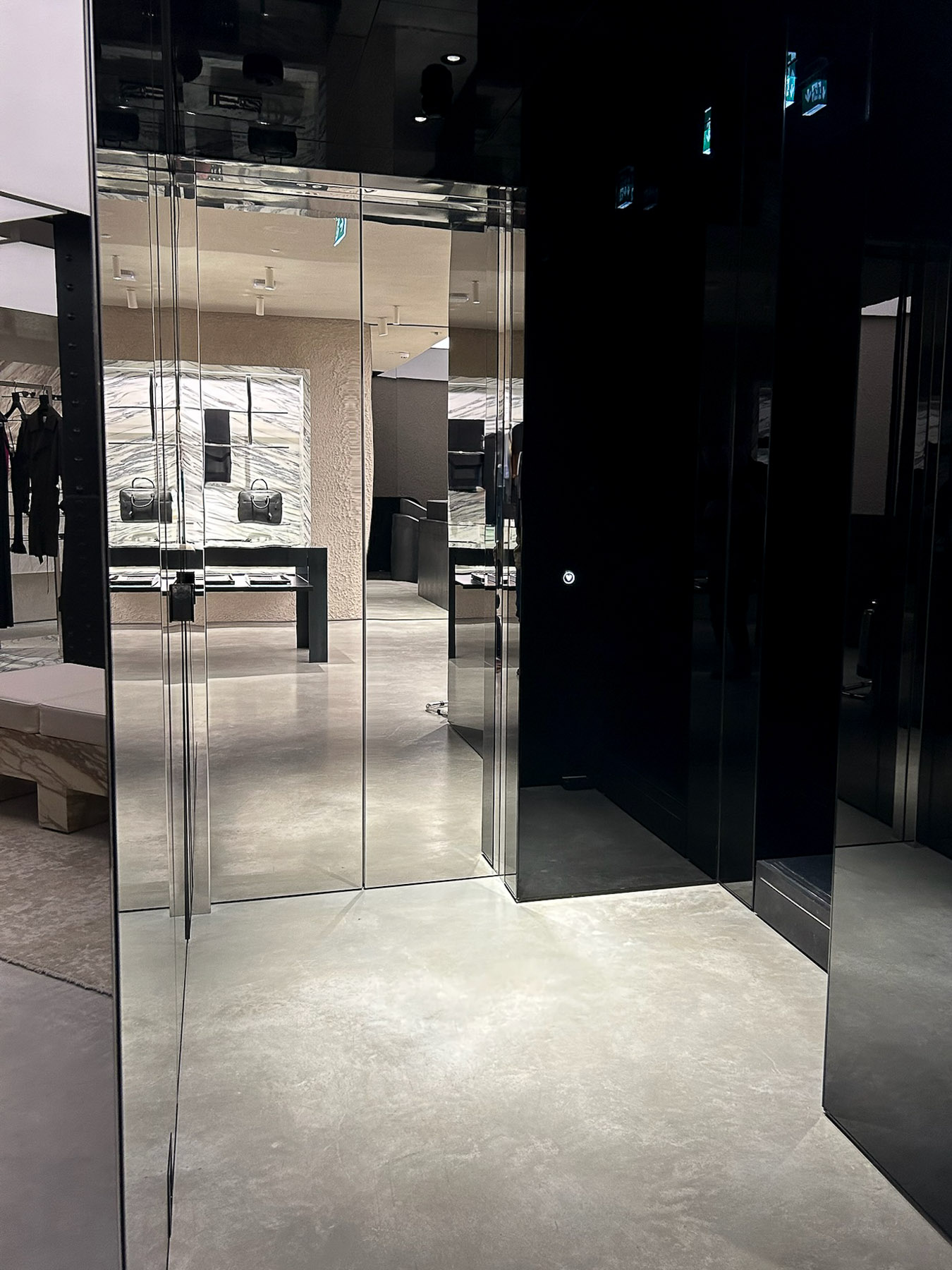 Modern retail store interior with polished concrete floor, glass and black reflective walls, and handbags displayed on illuminated marble-textured back panels.
