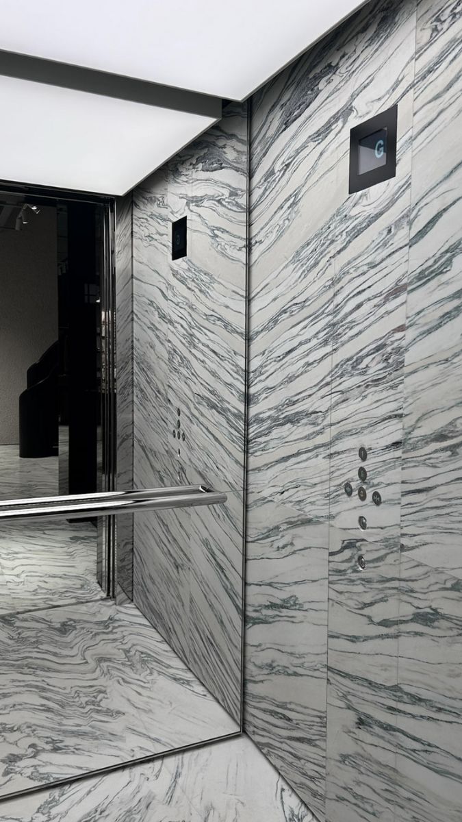 Interior of a modern elevator with white and gray marble walls and floor, stainless steel handrail, and illuminated ceiling panels.