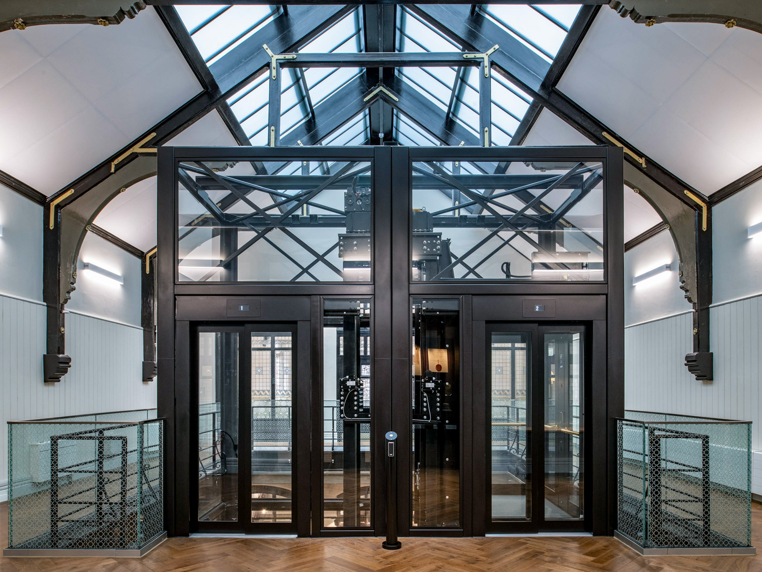Modern glass and black metal duplex passenger elevator inside a building with wooden floors and a glass ceiling.