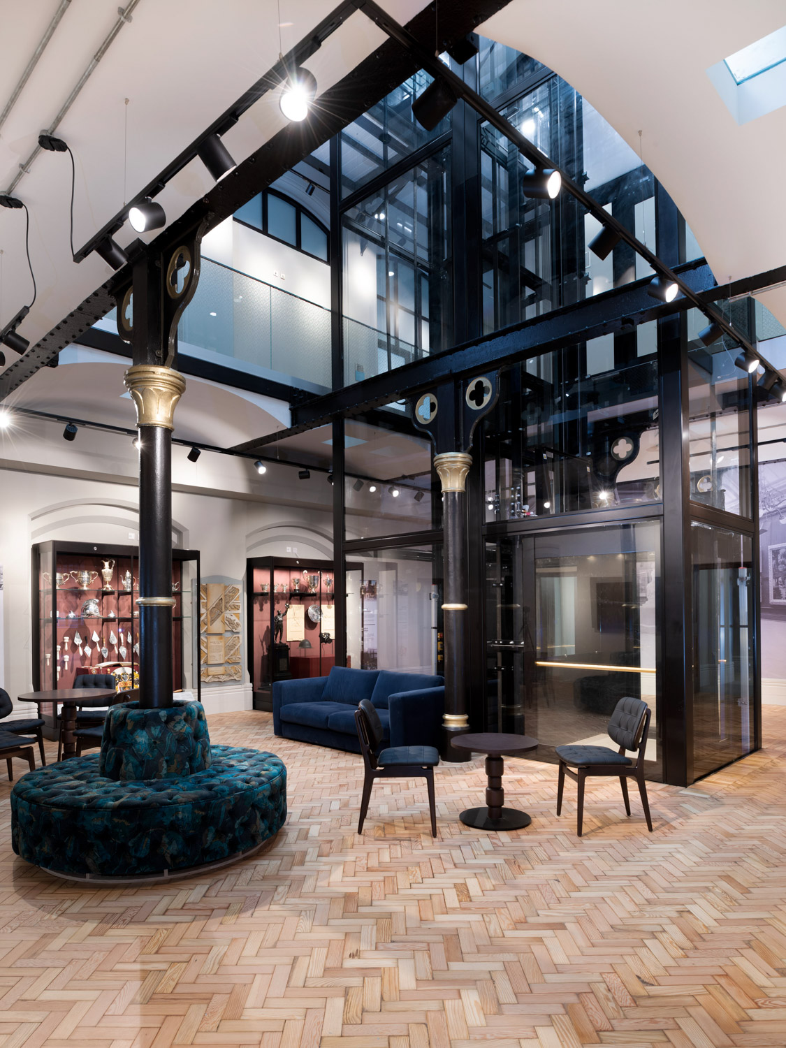 Modern gallery interior with polished wood herringbone floor, black and gold columns, glass elevator, blue velvet seating, and trophy displays.