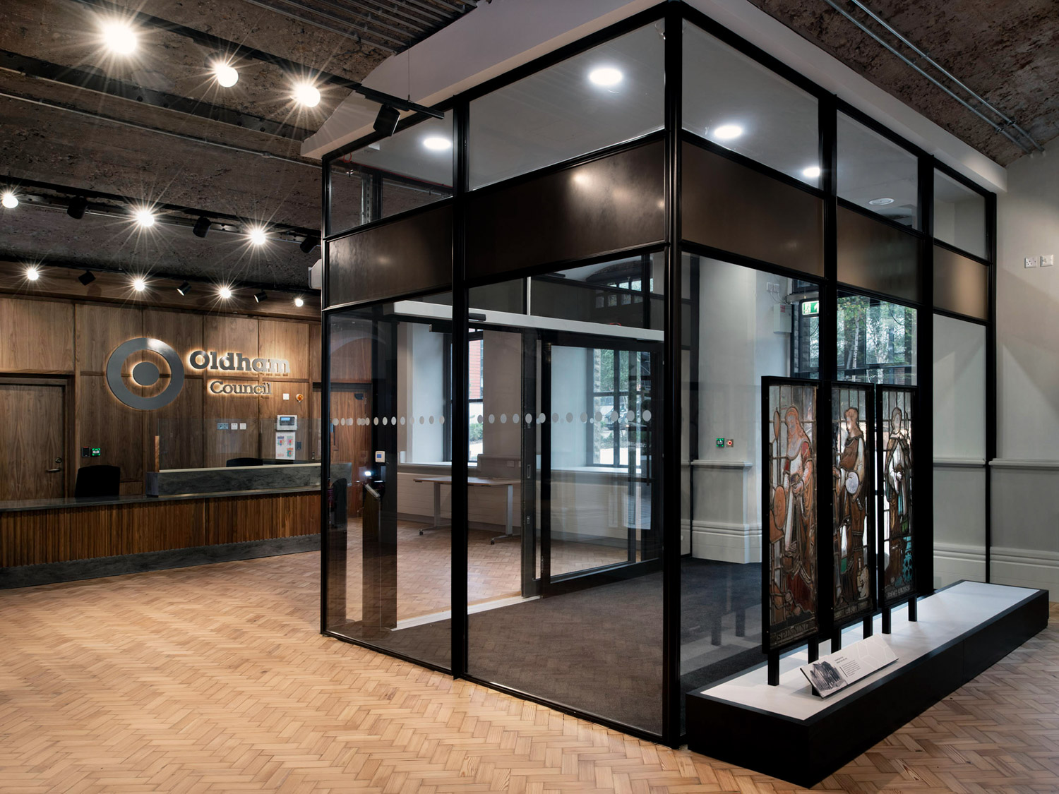 Modern reception area of Oldham Library with wood paneling, glass office enclosure, and stained glass artwork display.