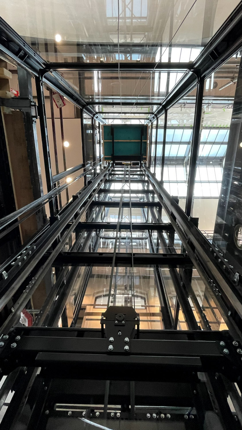 View looking up inside a modern glass and metal elevator shaft with cables and beams visible.