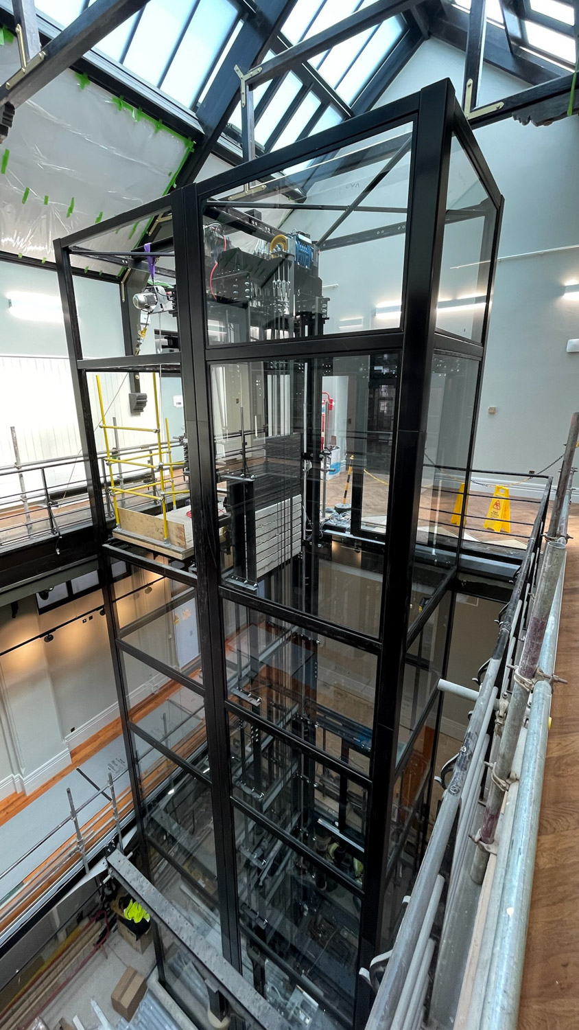 Modern glass and metal elevator shaft inside a building under construction or renovation with scaffolding around it.