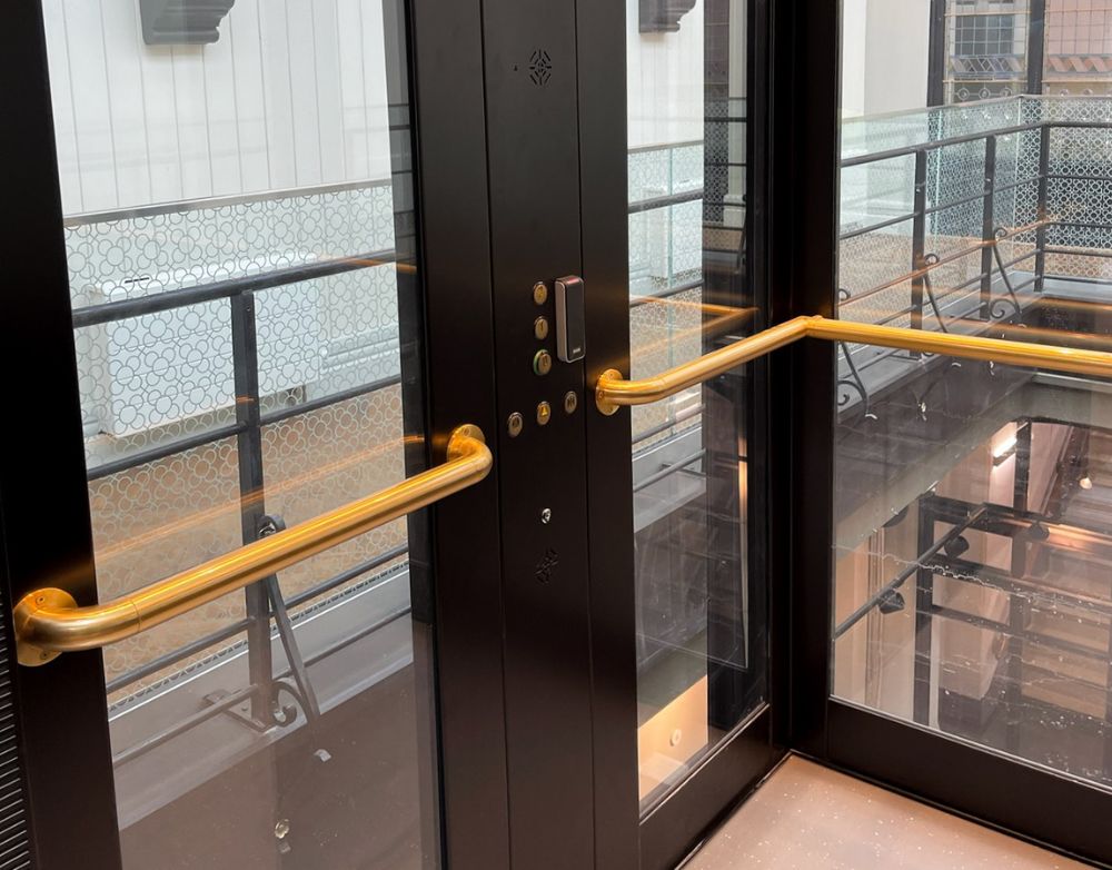 Interior view of a modern glass elevator with gold handrails and control buttons, overlooking a balcony with patterned glass panels.