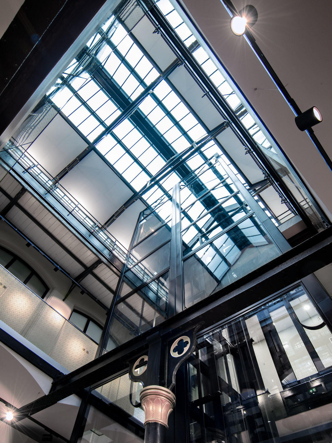 Modern glass elevator shaft inside a multi-story building with metal beams and a skylight ceiling.