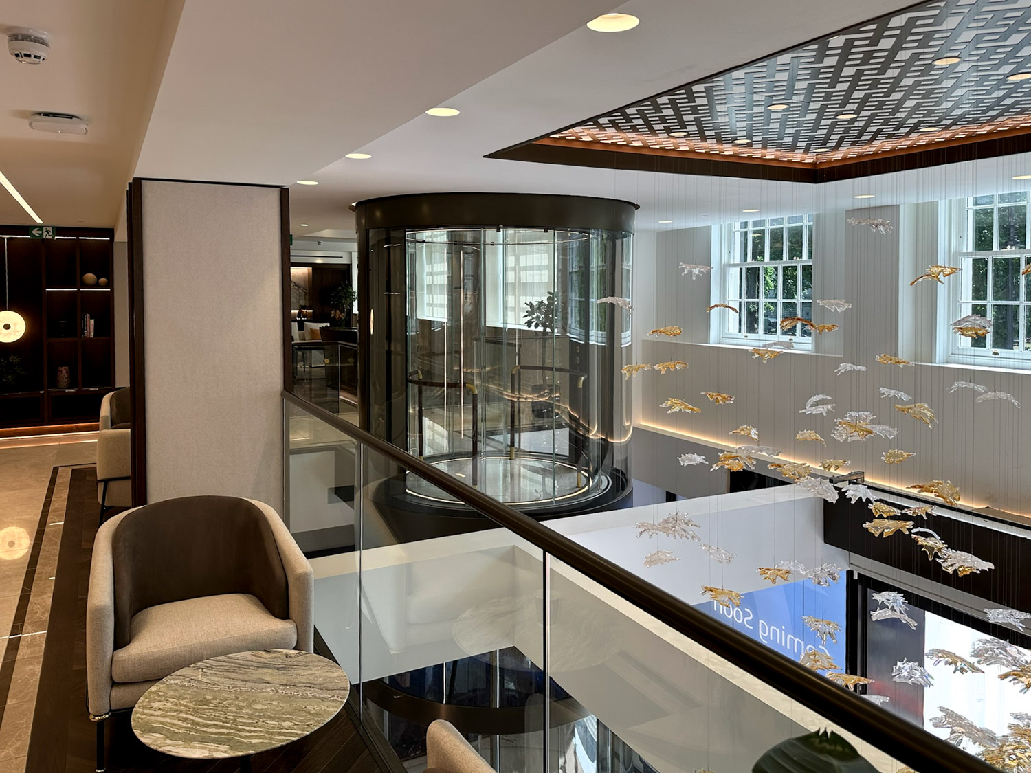 Modern interior with a round glass elevator, beige armchair, marble table, and hanging decorative leaves near large windows.