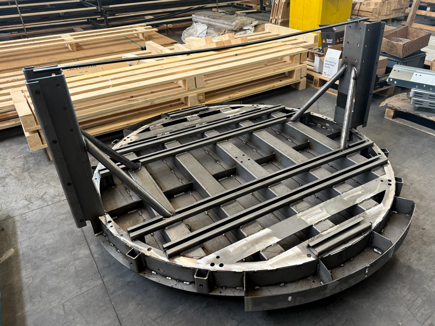 Large circular metal platform frame with welded joints, leaning on a warehouse floor surrounded by wooden planks and industrial materials.