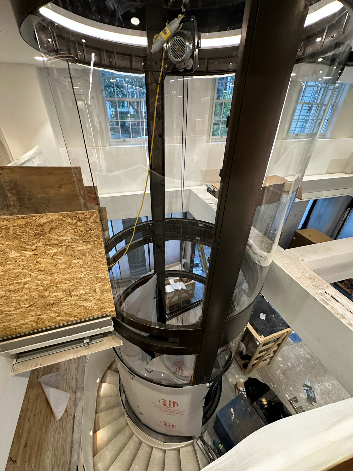 Glass cylindrical elevator shaft inside a building under construction with spiral staircase wrapping around it.