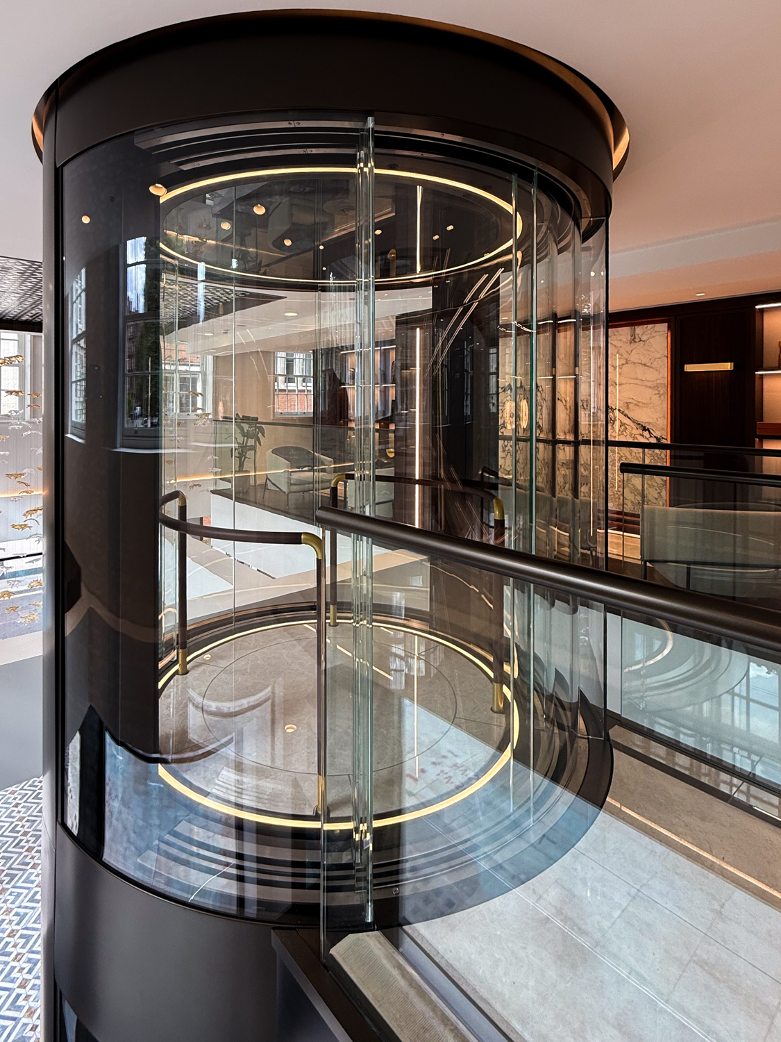 Modern glass cylindrical elevator inside a stylish building interior with reflective surfaces and circular LED lighting.