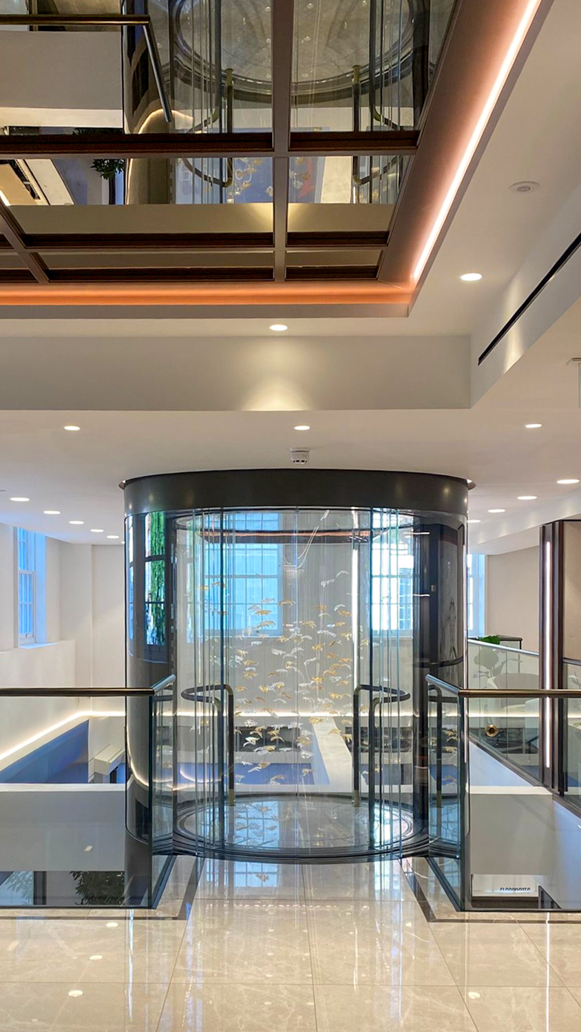 Modern glass cylindrical elevator inside a building lobby with illuminated 'FAB' logo and decorative ceiling installation.