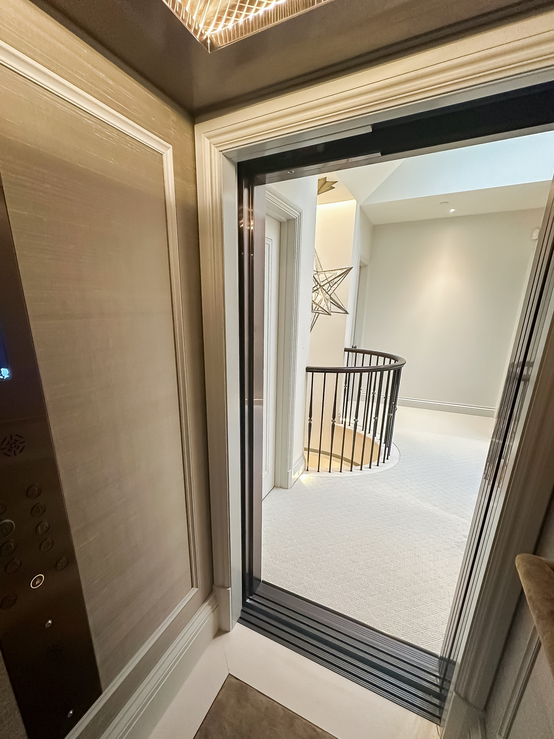 Interior view of a home elevator with its door open revealing a carpeted hallway and a curved staircase with black railings.