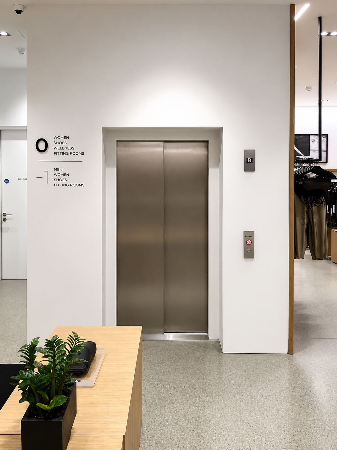 Closed stainless steel elevator doors inside a modern store with directional signs for women and men.