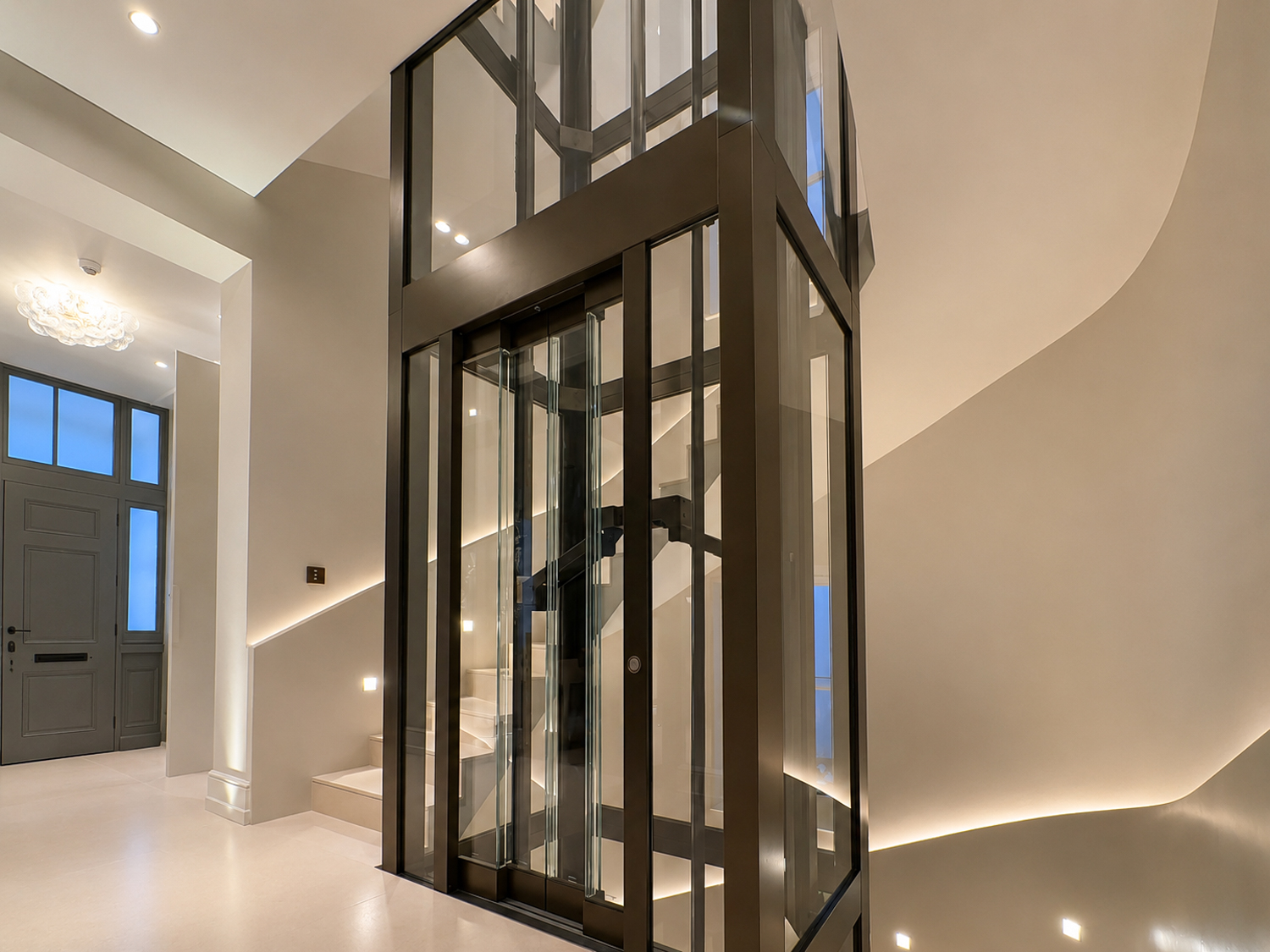 Glass passenger elevator in a well-lit home entrance with stairs and a gray door.