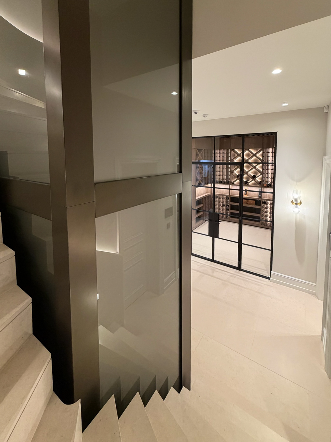 Modern interior with cream stairs leading past a glass lift to a wine cellar with black-framed doors.