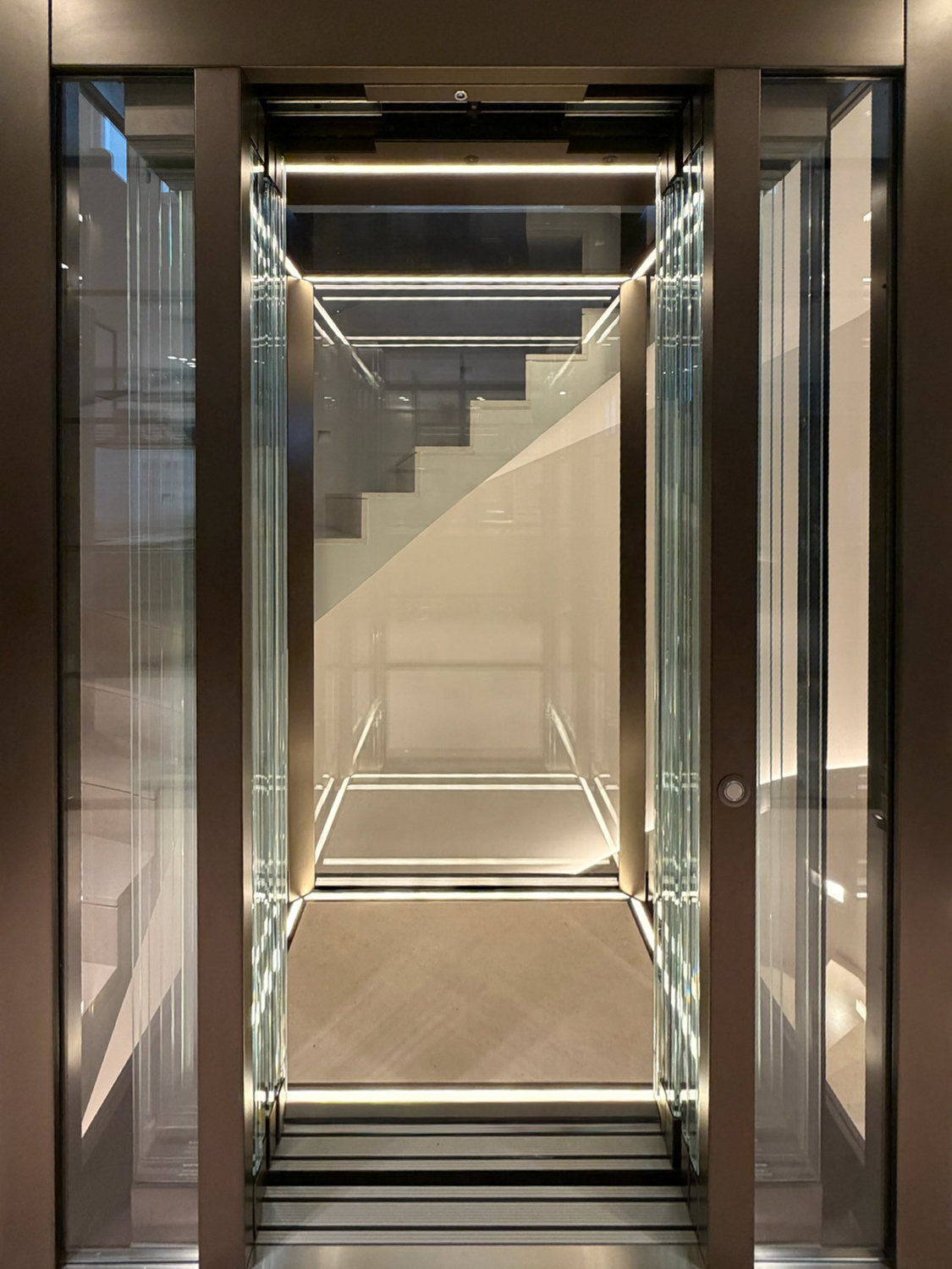 Modern glass lift cabin with illuminated edges highlighting a transparent staircase behind.