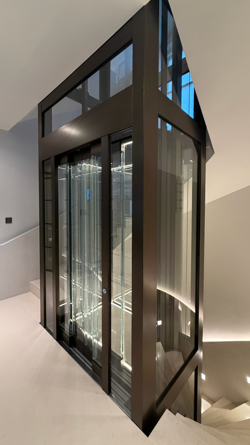 Modern glass passenger elevator with metal frame inside a clean, white staircase area.