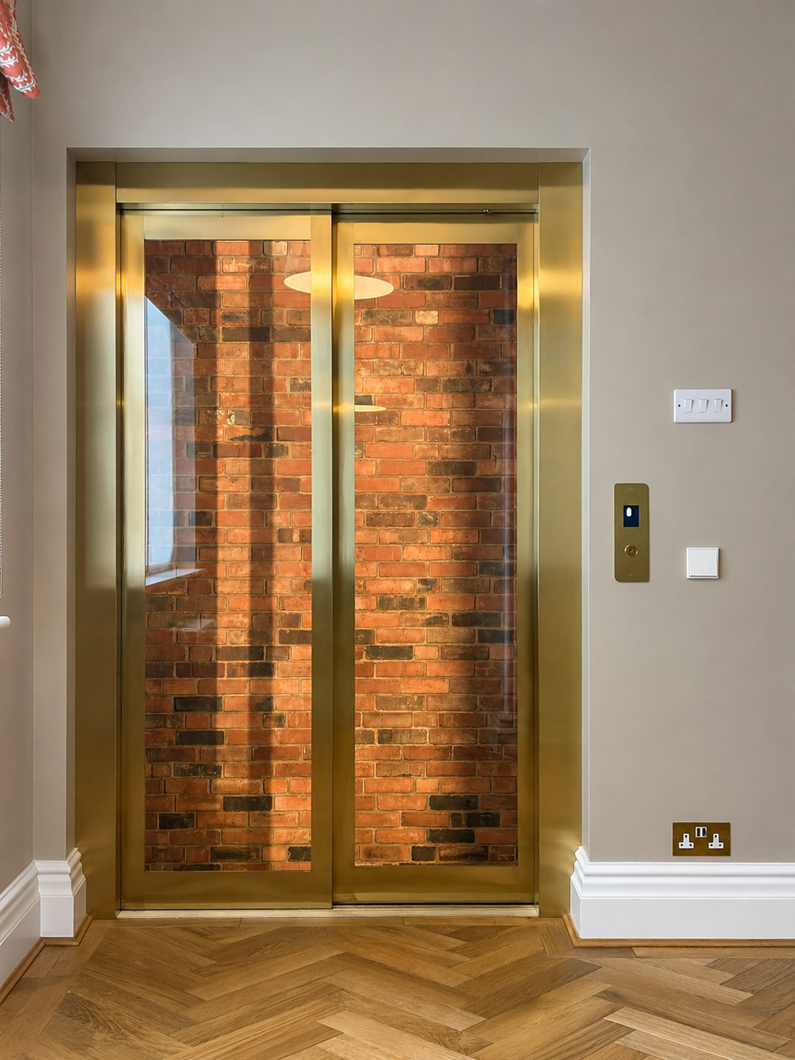 Gold framed glass elevator doors reflect brick wall and wooden herringbone floor inside a modern room.