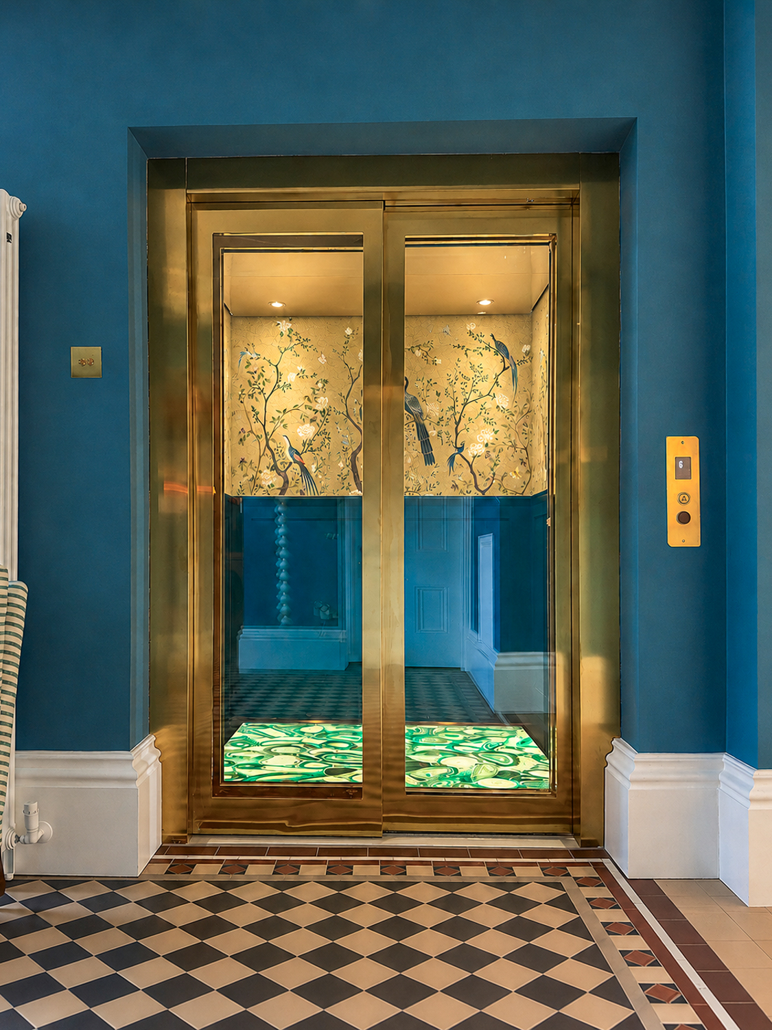 Gold-framed glass elevator doors by Premier Lift Group, in blue room with patterned floor and floral wallpaper inside elevator.