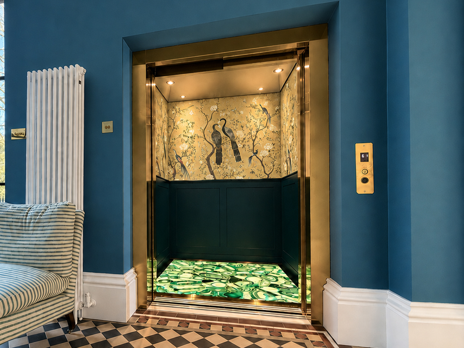 Modern home elevator with gold trim and bird mural, next to striped chaise lounge by large window.