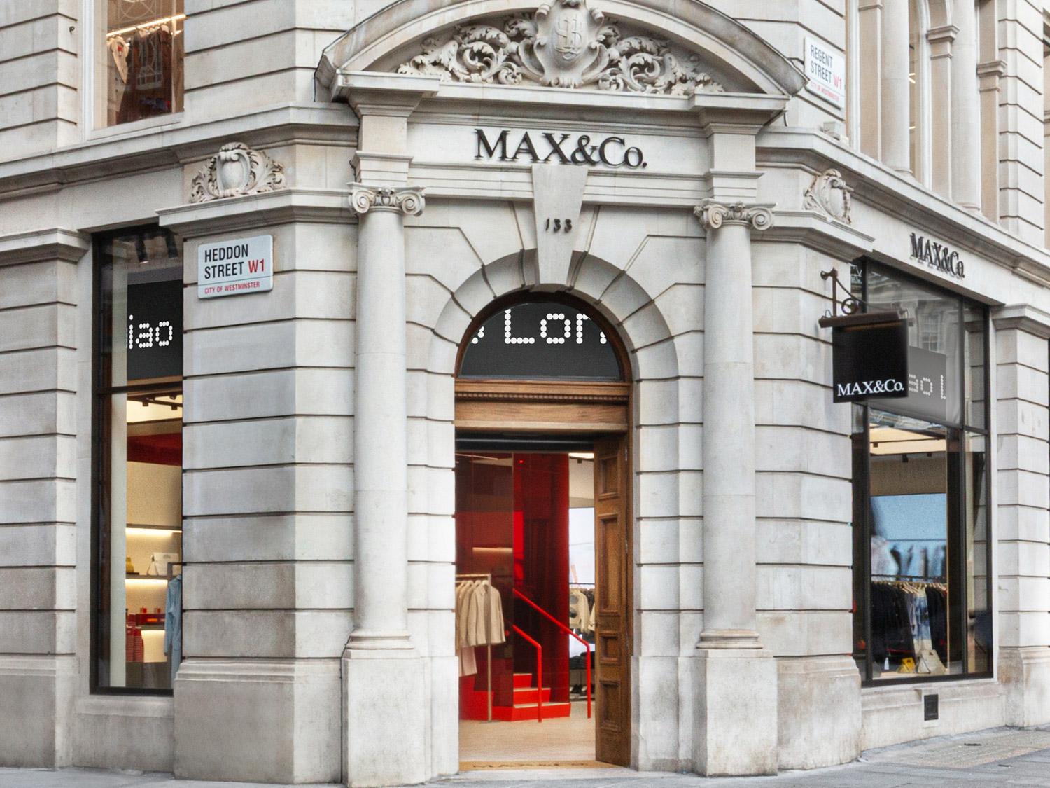 Stone building corner storefront with ornate architectural details housing a Max&Co. retail store; open wooden door reveals clothing racks and a red staircase inside; street signs indicate Heddon Street W1 and Regent Street W1 in the City of Westminster.