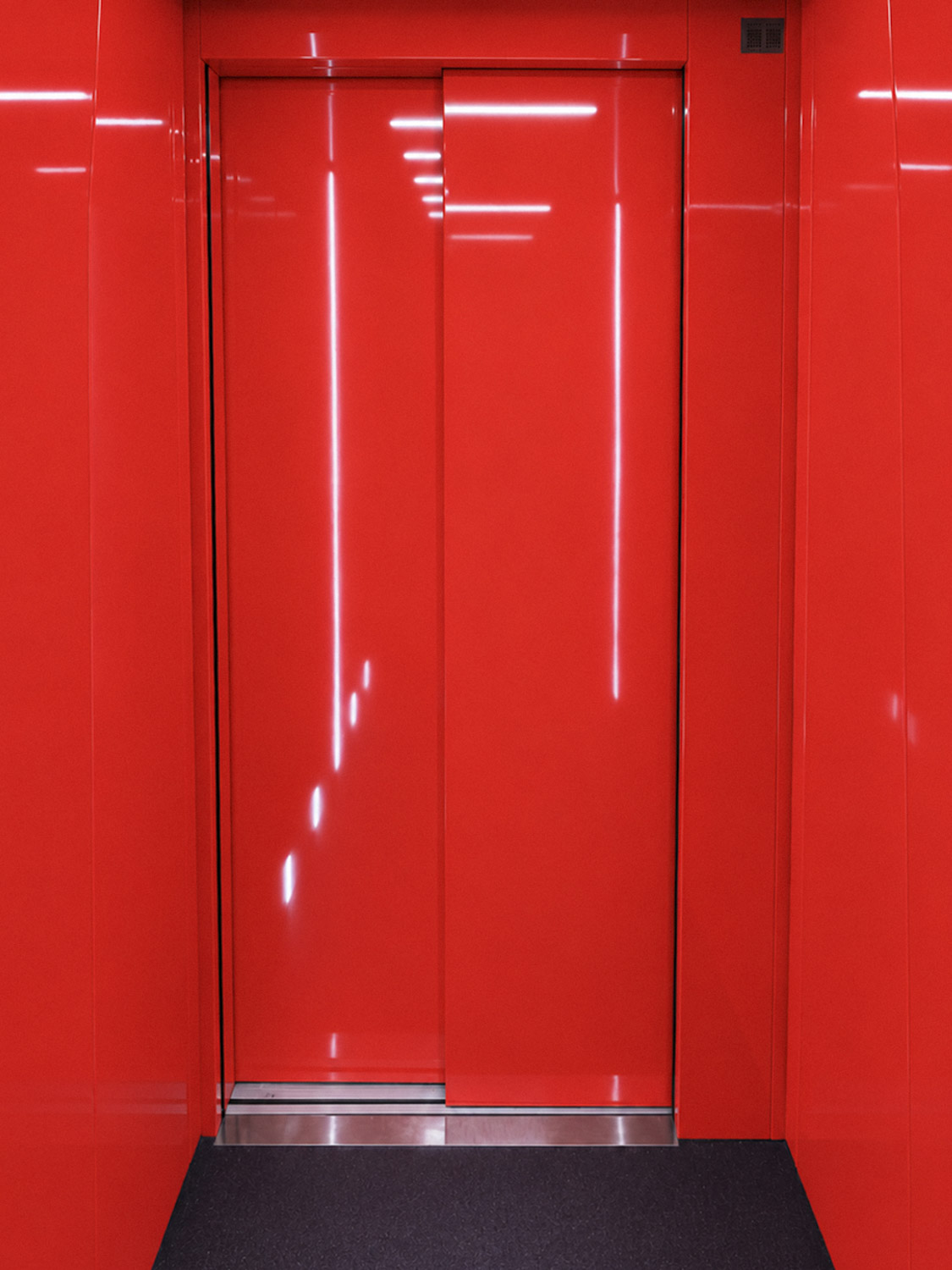 Closed bright red elevator doors with surrounding red wall panels and reflections of ceiling lights.