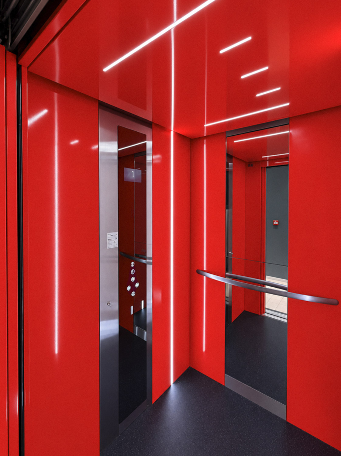 Interior of a modern elevator with bright red walls and ceiling, black floor, vertical and horizontal LED light strips, a metallic control panel with floor buttons, and a mirrored wall with a horizontal handrail reflecting the interior.
