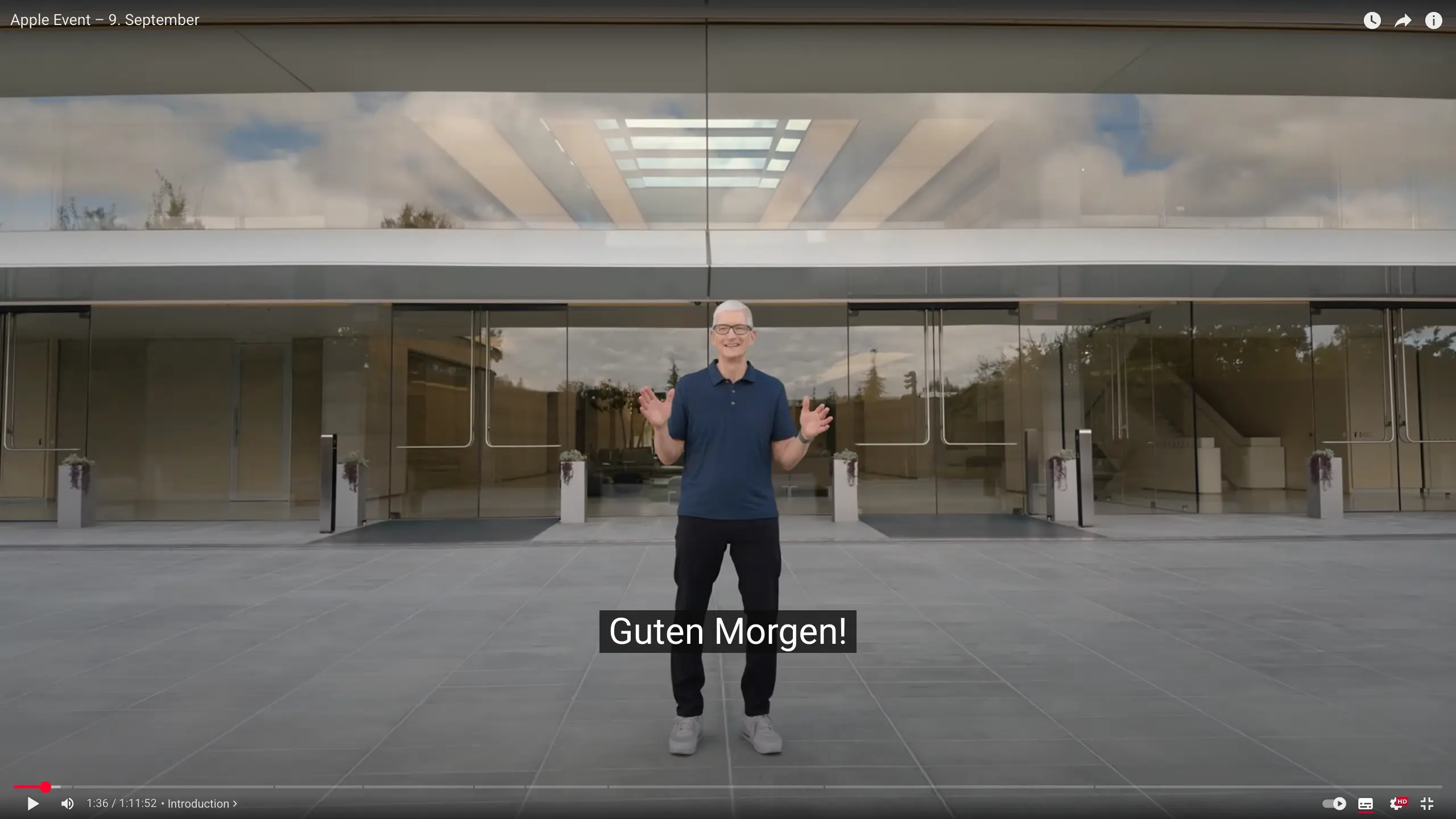 Die Bildschirmaufnahme zeigt Tim Cook, den CEO von Apple, wie er vor einem modernen, glasverkleideten Gebäude steht. Er trägt eine blaue Jeans, ein dunkelblaues Poloshirt und graue Turnschuhe. Seine Arme sind leicht erhoben, die Handflächen zeigen nach vorne. Auf dem Bildschirm ist ein Untertitel zu sehen, der die Worte "Guten Morgen!" anzeigt. Die Aufnahme stammt von einem Apple-Event, das am 9. September stattfand.