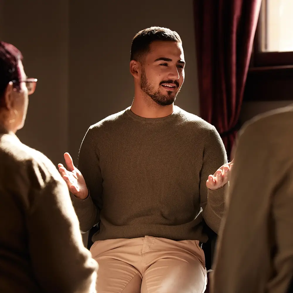  A group therapy session, in which a man is smiling and sharing with a mixed-gender group in a warm, safe therapy room.