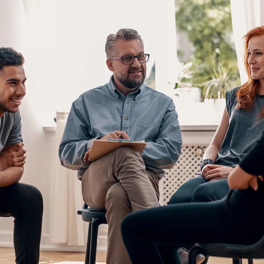 A mixed-gender group therapy session on a sunny day. The group is sitting by an open window, and one of the residents is speaking. Thanks to our rehab funding options, a diverse range of people can attend substance use rehab.
