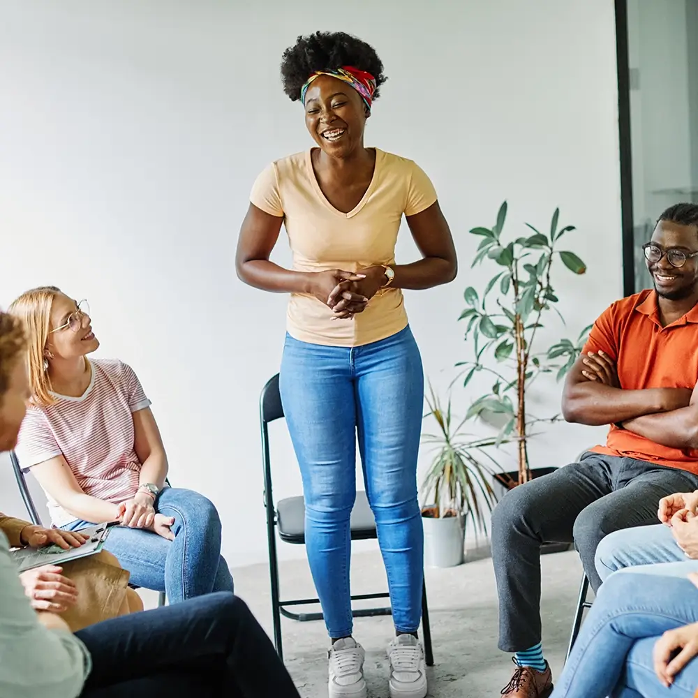 A smiling woman stands in the center of a seated support group, engaging warmly with others. The group members, diverse in appearance, are sitting in a circle in a bright, relaxed room with a potted plant in the background.