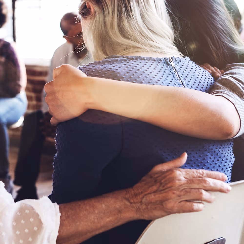 A therapy session, where a few of the residents have their arms over the shoulders of a female resident who’s sharing her story. 
